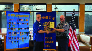 Two men shake hands as one presents an award plaque to the other, standing in front of banners and an American flag in a restaurant setting.