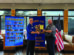 Two men shake hands as one presents an award plaque to the other, standing in front of banners and an American flag in a restaurant setting.