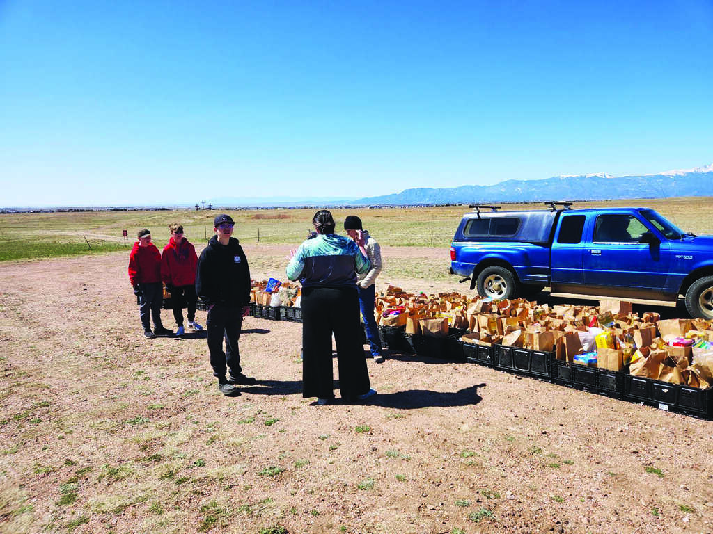 Several people stand near tables with bags of supplies next to a blue truck in an open field under a clear sky.