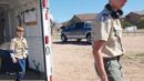 Two boys in scout uniforms, one stepping out of a trailer and the other walking on a dirt lot with houses and a parked truck in the background.