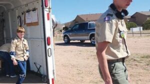 Two boys in scout uniforms, one stepping out of a trailer and the other walking on a dirt lot with houses and a parked truck in the background.