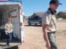 Two boys in scout uniforms, one stepping out of a trailer and the other walking on a dirt lot with houses and a parked truck in the background.