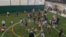 A group of adults and children participate in an activity on an indoor turf field; some gather objects on the ground while others look on.