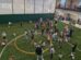 A group of adults and children participate in an activity on an indoor turf field; some gather objects on the ground while others look on.