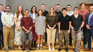 A group of fourteen people, including adults and teenagers, stand in two rows indoors, facing the camera and smiling.