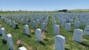 Rows of white headstones with small American flags placed beside them in a military cemetery under a clear blue sky.