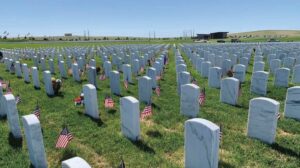 Rows of white headstones with small American flags placed beside them in a military cemetery under a clear blue sky.