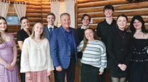 A group of nine young adults and one older man stand together indoors in front of log cabin walls, smiling at the camera.
