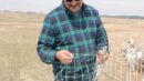 A man in a plaid shirt and cap inspects a section of wire fencing in a grassy field with sheep in the background.