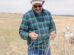 A man in a plaid shirt and cap inspects a section of wire fencing in a grassy field with sheep in the background.