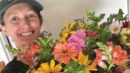 A person wearing a green cap smiles while holding a large, colorful bouquet of various flowers in front of a light background.