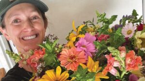 A person wearing a green cap smiles while holding a large, colorful bouquet of various flowers in front of a light background.