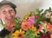 A person wearing a green cap smiles while holding a large, colorful bouquet of various flowers in front of a light background.