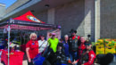 A group of people pose beside a blue motorcycle in front of an American Legion Auxiliary tent outside a building.