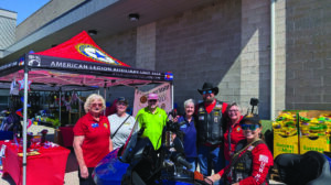 A group of people pose beside a blue motorcycle in front of an American Legion Auxiliary tent outside a building.
