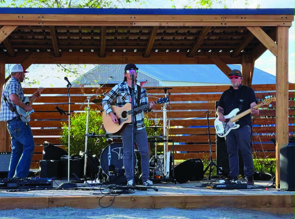 Four musicians perform on an outdoor wooden stage, playing guitar, bass, and drums under a pergola, with microphones and music equipment set up around them.