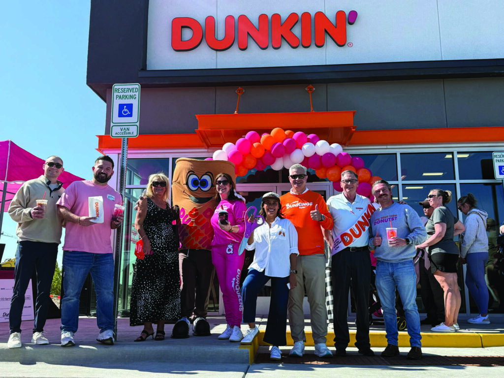 A group of people stand in front of a Dunkin’ store, holding beverages, with a Dunkin’ mascot and balloon decorations visible in the background.