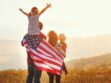 A family stands in a field at sunset, with one child on an adult's shoulders and an American flag draped around them.