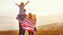 A family stands in a field at sunset, with one child on an adult's shoulders and an American flag draped around them.