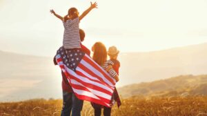 A family stands in a field at sunset, with one child on an adult's shoulders and an American flag draped around them.
