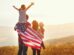 A family stands in a field at sunset, with one child on an adult's shoulders and an American flag draped around them.