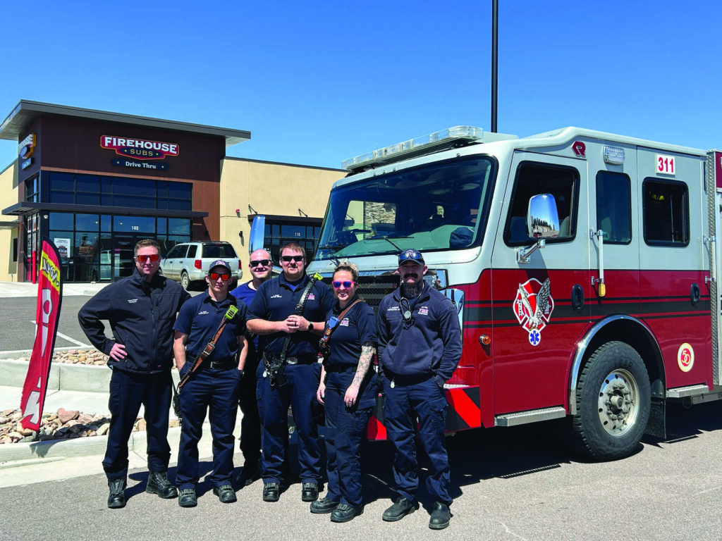 Six firefighters in uniform stand in front of a fire truck parked near a Firehouse Subs restaurant on a sunny day.
