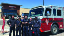 Six firefighters in uniform stand in front of a fire truck parked near a Firehouse Subs restaurant on a sunny day.
