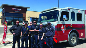 Six firefighters in uniform stand in front of a fire truck parked near a Firehouse Subs restaurant on a sunny day.