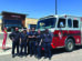 Six firefighters in uniform stand in front of a fire truck parked near a Firehouse Subs restaurant on a sunny day.