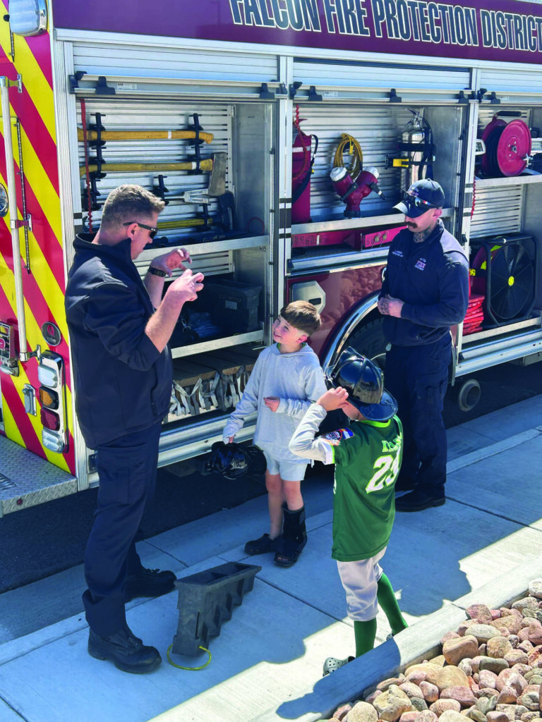 Two firefighters stand by a fire truck showing equipment to two young boys, one of whom is wearing a green baseball uniform.