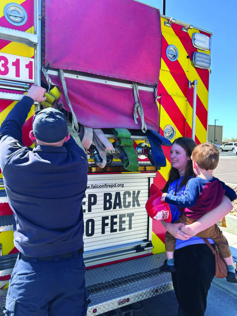 A firefighter shows a fire truck’s equipment to a woman holding a child in a parking lot on a sunny day.