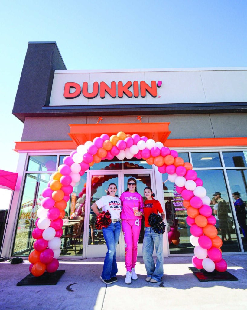 Three people stand under a pink, orange, and white balloon arch in front of a Dunkin' store on a sunny day.