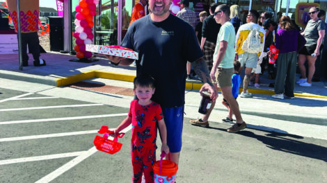A man and a child stand in front of a Dunkin' store entrance decorated with balloons, holding Dunkin' merchandise, with a crowd and cars in the background.