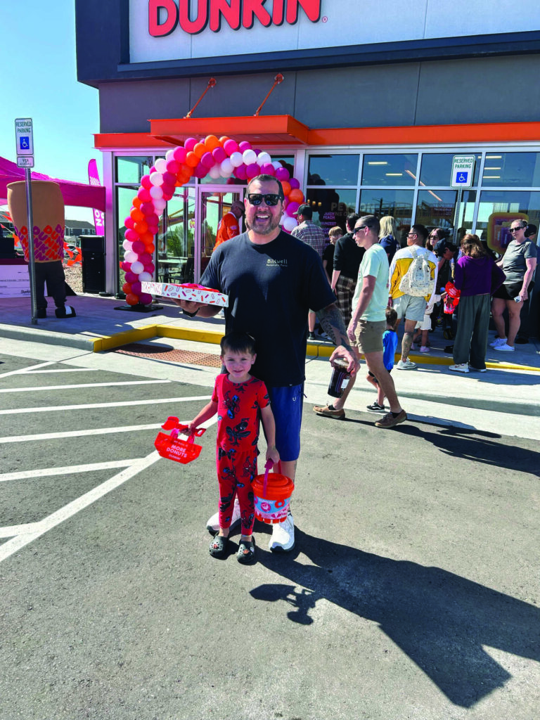 A man and a child stand in front of a Dunkin' store entrance decorated with balloons, holding Dunkin' merchandise, with a crowd and cars in the background.