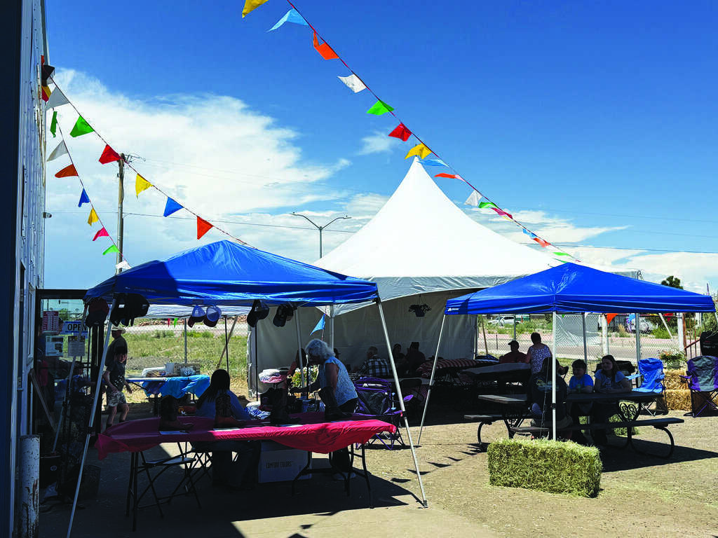 Outdoor event with people sitting under blue and white tents, colorful pennant flags overhead, and tables set up on a sunny day.