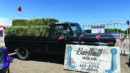 A black pickup truck loaded with hay bales is parked behind a large Damman Hay Co. sign at an outdoor event, with small American flags on the truck.