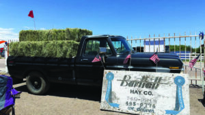 A black pickup truck loaded with hay bales is parked behind a large Damman Hay Co. sign at an outdoor event, with small American flags on the truck.