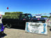 A black pickup truck loaded with hay bales is parked behind a large Damman Hay Co. sign at an outdoor event, with small American flags on the truck.