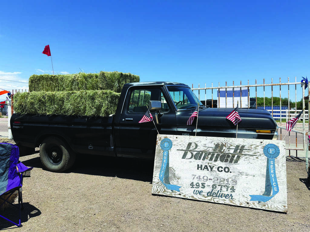A black pickup truck loaded with hay bales is parked behind a large Damman Hay Co. sign at an outdoor event, with small American flags on the truck.