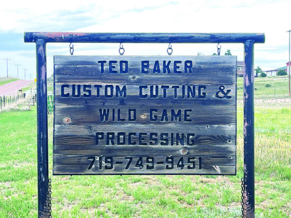 Wooden sign reads: "Ted Baker Custom Cutting & Wild Game Processing 719-749-9451." The sign hangs outdoors with grass and a road in the background.