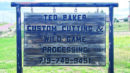 Wooden sign reads: "Ted Baker Custom Cutting & Wild Game Processing 719-749-9451." The sign hangs outdoors with grass and a road in the background.