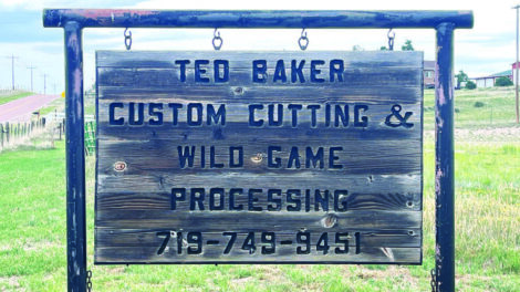 Wooden sign reads: "Ted Baker Custom Cutting & Wild Game Processing 719-749-9451." The sign hangs outdoors with grass and a road in the background.