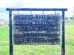 Wooden sign reads: "Ted Baker Custom Cutting & Wild Game Processing 719-749-9451." The sign hangs outdoors with grass and a road in the background.