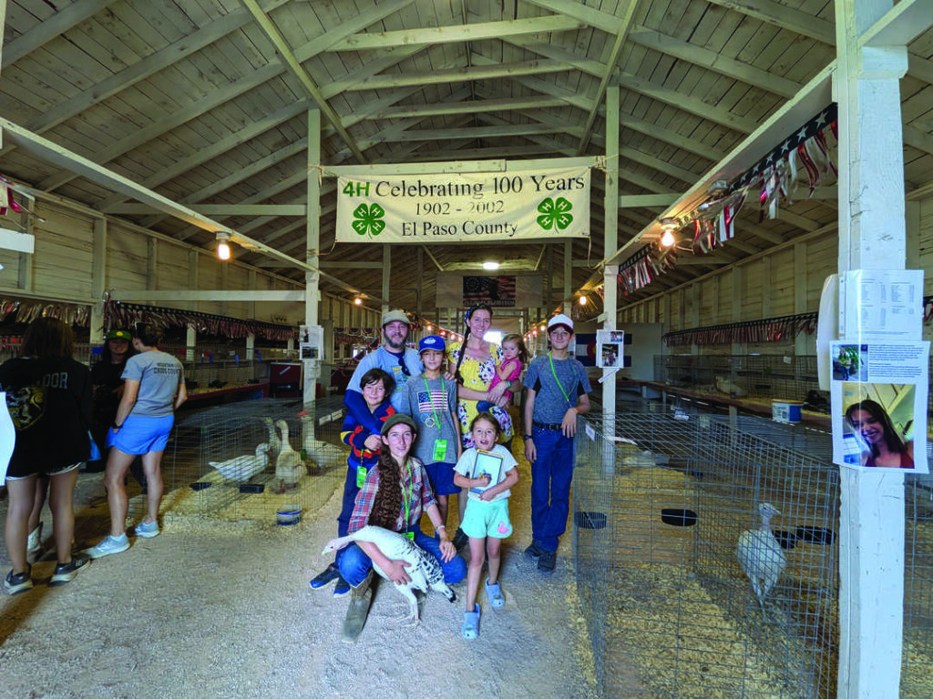 A group of people, including children, pose in a barn with animals under a sign reading "4H Celebrating 100 Years 1902-2002 El Paso County.