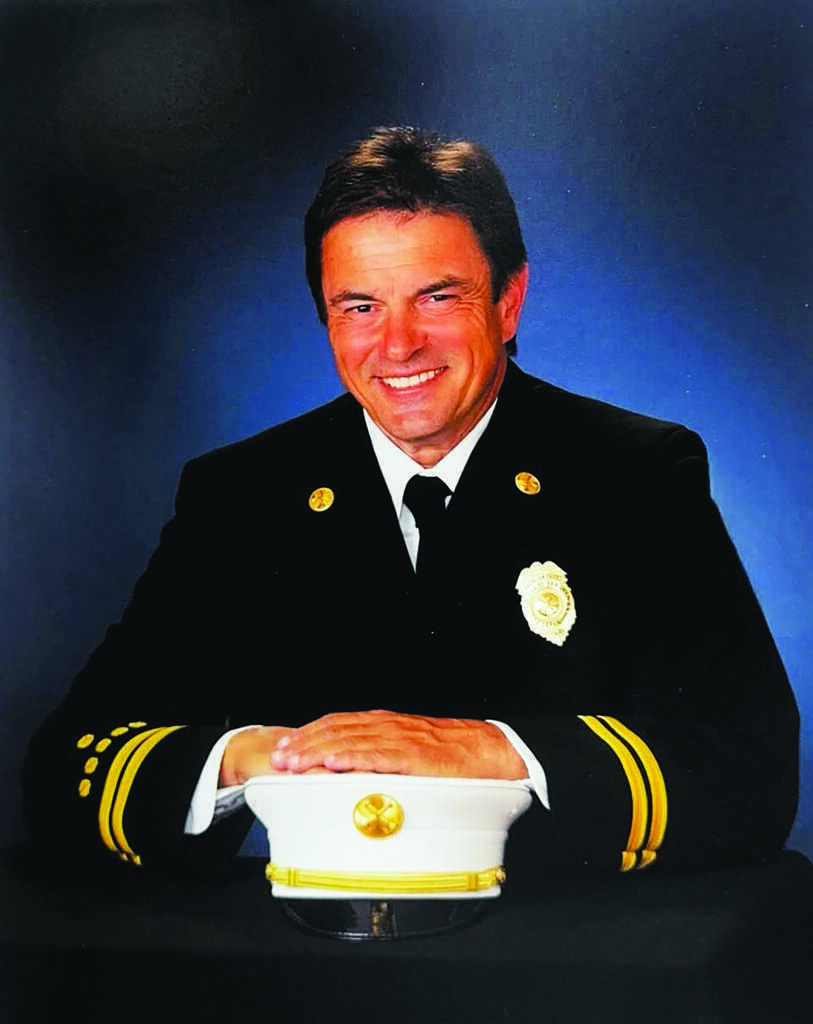 Man in a formal fire chief uniform sits at a table with his white fire chief hat in front of him, posing for a professional portrait against a blue background.