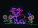 Colorful LED lights form the image of a cartoon cow standing on grass with flowers, set against a black background.