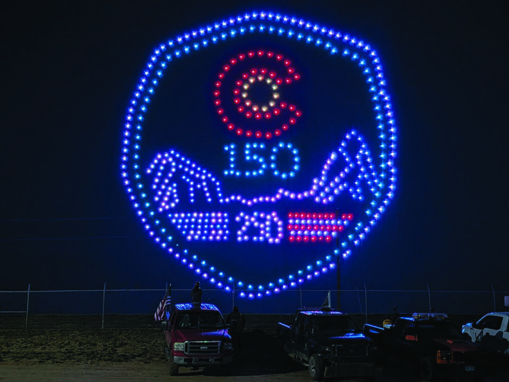 A formation of drones creates an illuminated Colorado emblem with "150" and "2023" above parked vehicles at night.