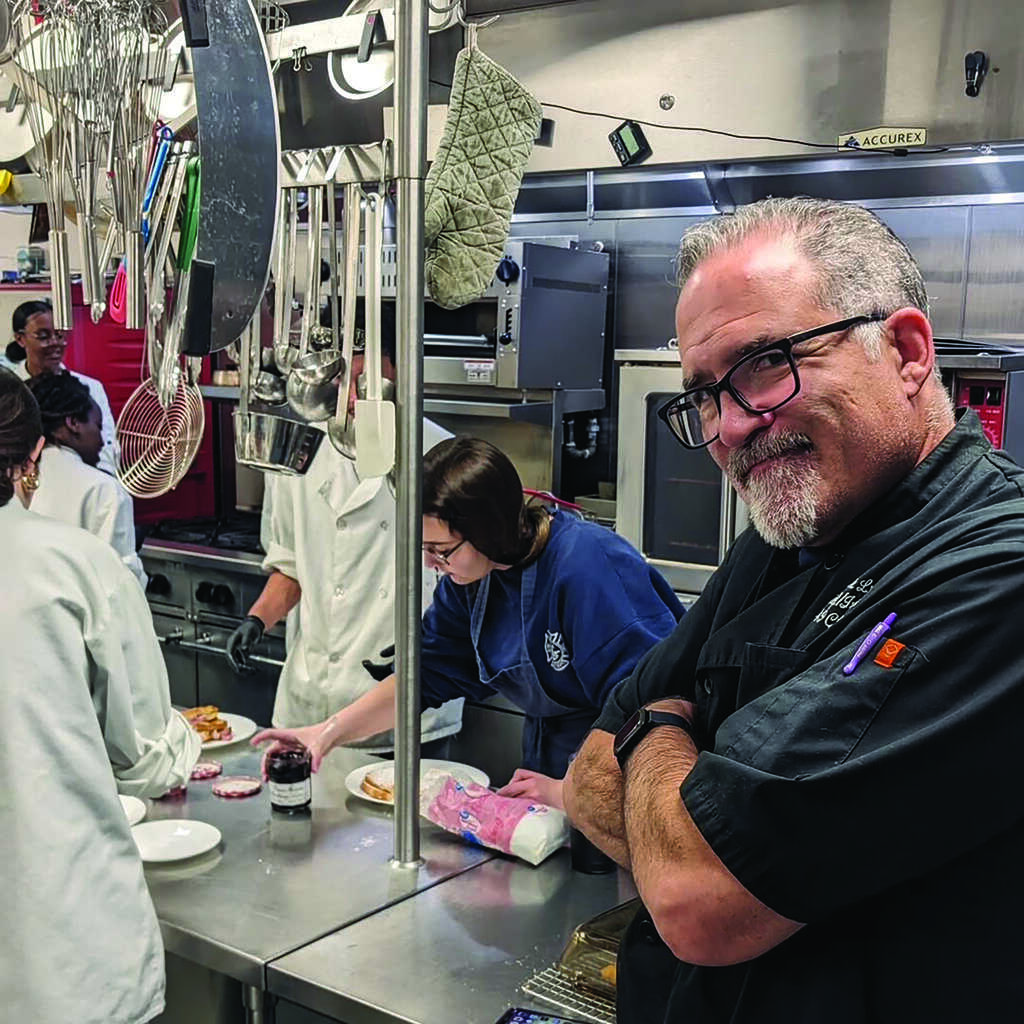 A chef stands with arms crossed in a commercial kitchen while several people in chef coats work at the counter behind him, preparing food.