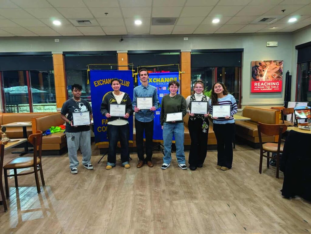 Six people stand indoors holding certificates, posing for a group photo in front of Exchange Club banners in a well-lit room with tables and chairs.