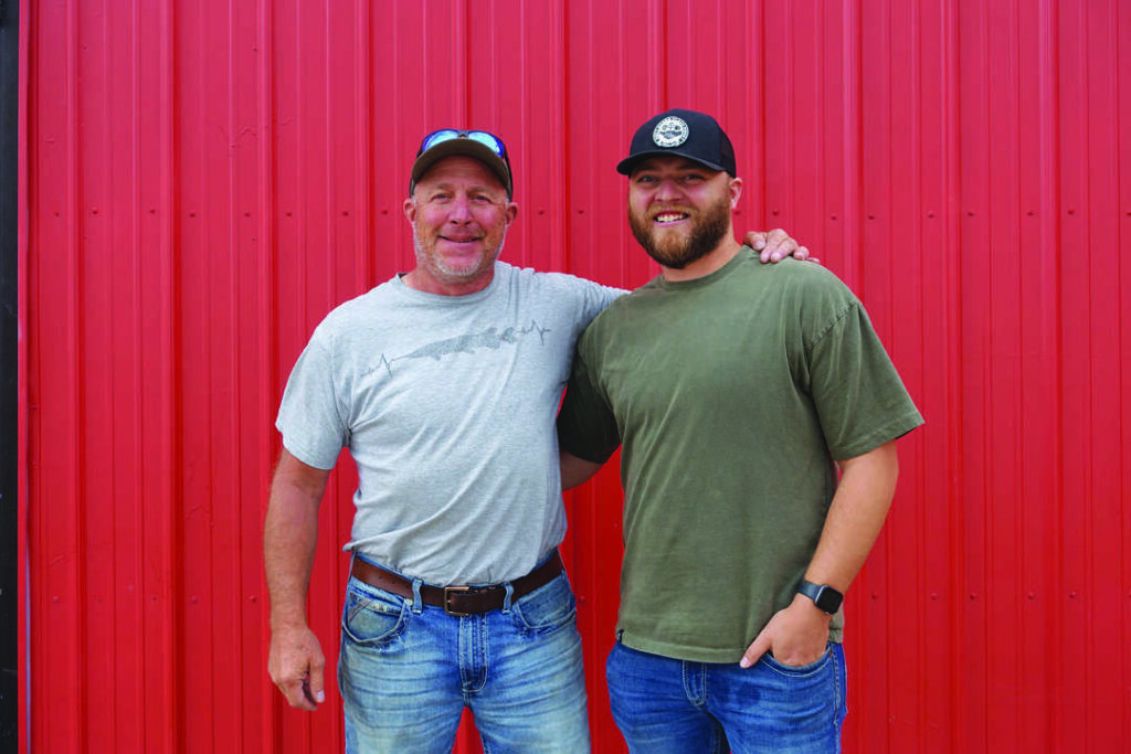 Two men standing side by side in casual clothes, smiling in front of a red metal wall.